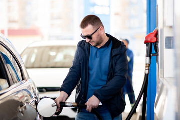 A man with a blue jacket and dark glasses at a gas station. He fills up the car. Petrol. Lifestile.
