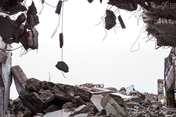 A hole in the body of a building with a pile of construction debris and concrete fragments hanging on the rebar against a uniform gray sky. Background - obrazy, fototapety, plakaty