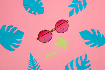 Fototapeta na wymiar sunglasses on a pink background under a paper palm tree around tropical blue leaves, creative summer concept
