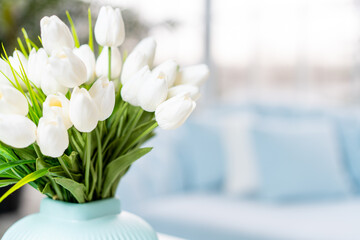 White tulip flowers in vase standing coffee table with blurred interior background.