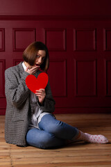 Surprised short-haired woman in jacket sitting on floor holding heart shape in front of her