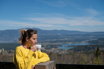 Fototapeta na wymiar Young woman drinking coffee with enjoyment outside on a terrace with a beautiful view of a lake