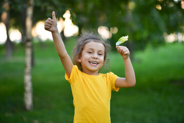 A little cute girl in a yellow T-shirt against the background of green trees.