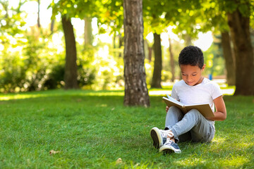 Little African-American boy reading book on grass in park