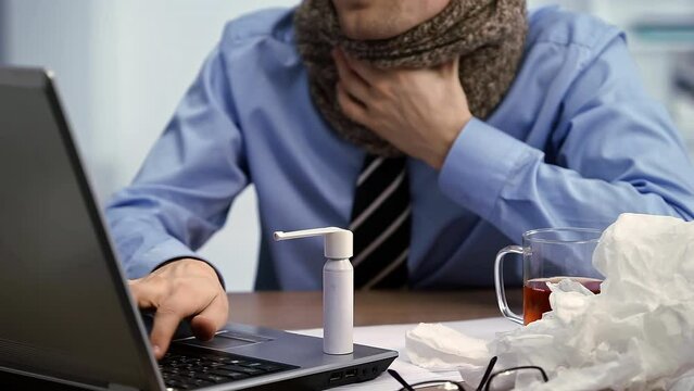 Throat Pain. Sick worker using spray for throat. Young man working in office suffering virus of flu, works on his laptop in the office.