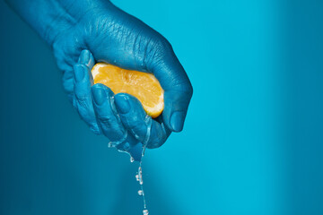 Close-up shot of male hand painted in blue squeezing ripe half of lemon, its juice dripping through...