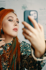 Adorable woman takes a selfie on the front camera of a modern smartphone. Female blogger shoots herself for a blog about fashion and makeup. Red-haired girl with a ethnic slavic crown on her head.