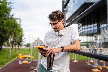 One caucasian man taking a brake during outdoor training in the park outdoor gym using mobile phone texting or reading typing resting on the bars with supplement shaker in hand happy smile copy space