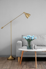 Vase with bouquet of tulips on table near sofa and modern floor lamp near light wall