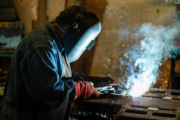 Welder Industrial works in an old workshop, holds a welding machine in his hand and welds metal structures at a factory.