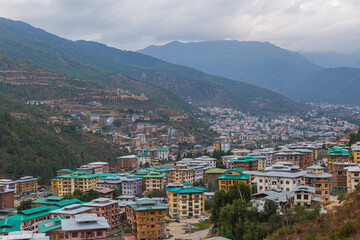 Fototapeta na wymiar Thimphu, Bhutan - October 26, 2021: Aerial view cityscape of Bhutan capitol city. Top view with dramatic cloudy sky over the town. Largest city in Bhutan in a mountain valley. Houses with green roof