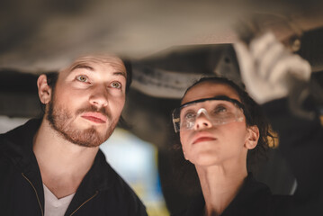 Young male and female car mechanic wearing safety glasses and hand gloves standing under car in garage inspecting and doing maintenance work for car