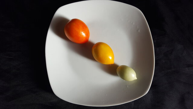 Three freshly picked tomatoes, small, medium and slightly larger are lined diagonally on a white plate. The tomatoes are red or ripe, yellow or half ripe and green.   