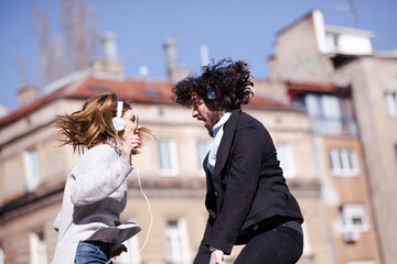 Young business colleagues dancing together with headphones after work. Happy friends jumping and fooling around outdoors