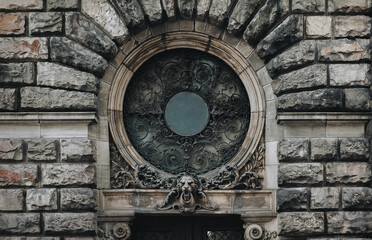 A round window with an openwork forged metal grill on the facade of an old house with stucco and decorative relief elements and a lion's head above the doorway. Lviv, Ukraine.
