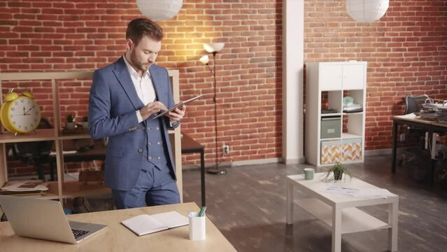 A man in the office walks in circles and looks at the computer tablet, then goes to the laptop and types something, then moves away and continues to do something on the tablet. Slow motion 4k footage