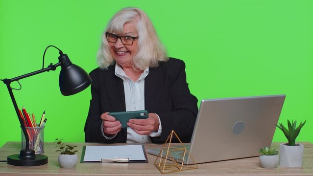 Worried funny senior businesswoman sits at workplace avoid working and enthusiastically playing racing or shooter video games at office desk table. Elderly woman on green chroma key studio background
