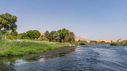 Fototapeta na wymiar The bed of the Nile River bends. Green vegetation grows on the banks. At the top of a sand dune, against a clear sky, the Aga Khan mausoleum is visible. Ripples on the blue water. Egypt. Aswan