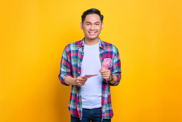 Smiling young Asian man in plaid shirt holding money banknotes and looking at camera isolated on yellow background