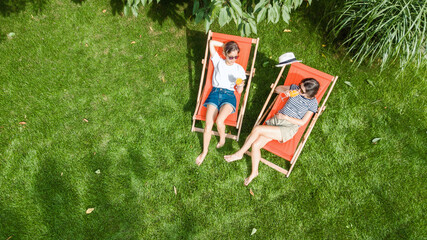 Young girls relax in summer garden in sunbed deckchairs on grass, women friends have fun outdoors...