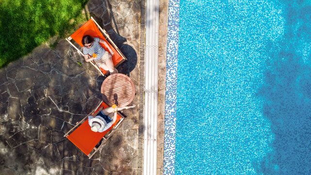 Young girls relax near swimming pool in sunbed deckchairs, women friends relax on tropical vacation in hotel resort, aerial drone view from above