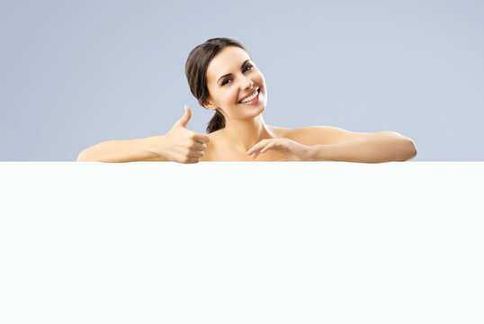 Excited smiling brunette woman with naked shoulders, undress, showing thump up hand sign gesture, standing behind white banner mock up signboard with copy space area, over grey color background.