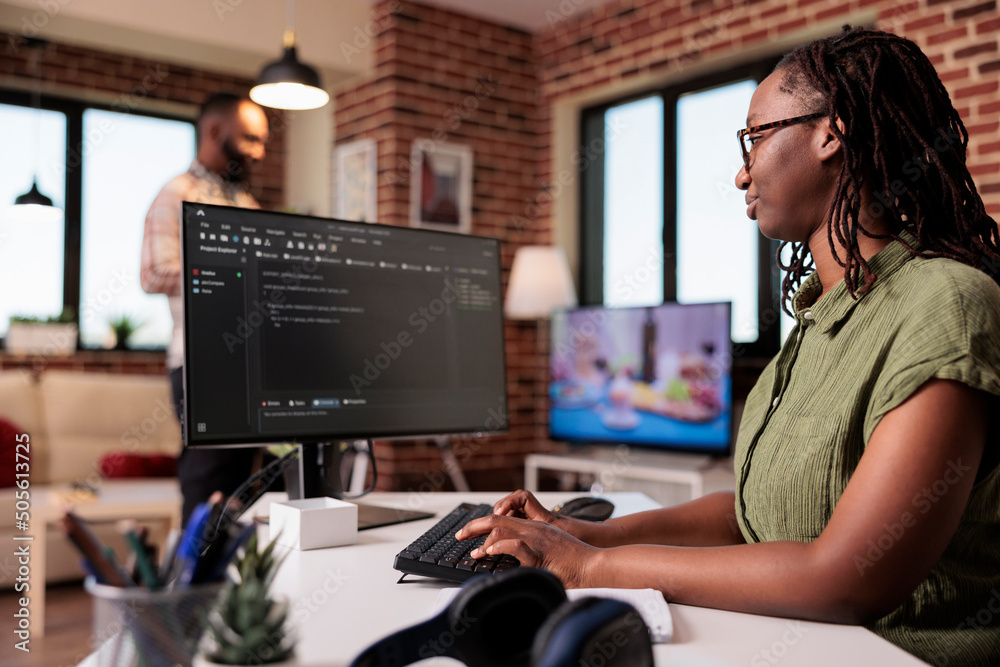 Poster woman programmer working remote writing code using computer while boyfriend is relaxing watching tv.