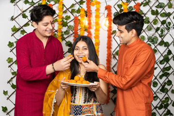 Portrait of happy indian young kids or bothers and sister wearing traditional cloths having fun eating laddu or laddoo sweets celebrating festival like diwali or rakshabandhan,