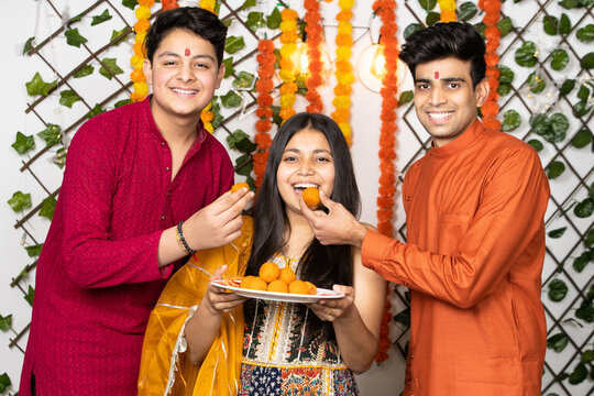Portrait of happy indian young kids or bothers and sister wearing traditional cloths having fun eating laddu or laddoo sweets celebrating festival like diwali or rakshabandhan,
