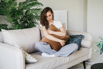 Obraz premium A sad woman sits on a sofa at home hugging a pillow. 