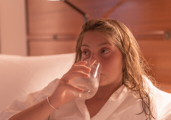 A young caucasian girl drinks a glass water on the bed. 