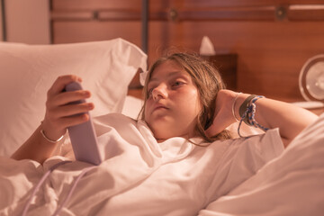 A young girl wearing a white bathrobe lies down on the bed playing with her phone. 