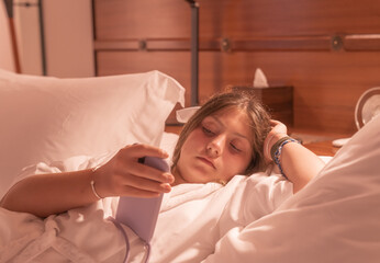 A young girl wearing a white bathrobe lies down on the bed playing with her phone. 