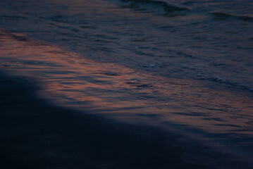 Surf of water on sunset with dark colorful pink and indigo blue reflection in water, black diagonal coastline, closeup. Water texture, background.
