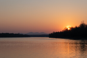 Landscape of river and mountantain before sunset, rippling water and distant mountain in haze with sun setting behind forest on clear sky, calming fair weather in nature