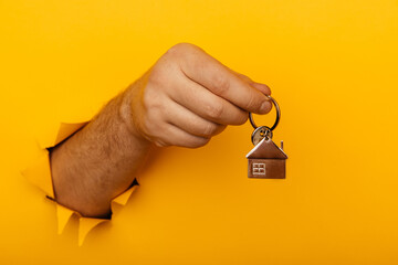 Male hand with metal house and key through a hole in yellow background. House sale and rent concept