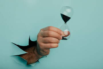 Male hand holds hpurglass in torn hole of blue background