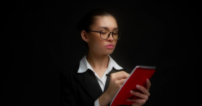 Dissatisfied business woman with glasses does not like what is written in a red notebook, she crosses everything out. Beautiful brunette in business clothes, isolated on a black background