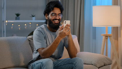 Happy arabian indian male millennial man holding modern smartphone texting message at home sitting...
