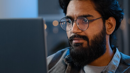 Close-up bearded male face reflection light from laptop monitor in glasses. Busy serious focused Arab Indian man looking at computer working freelance in evening playing game at night surfing in net