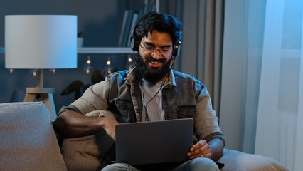Arabian indian funny happy bearded man wears wireless headphones having fun listening music with laptop at home sitting couch evening late time darkness room relaxing singing song enjoy audio app
