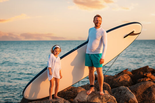 Summer activity vacations. Happy father and daughter standing with sup board on big beach rocks. Sunset sky and ocean at the background. Surfing