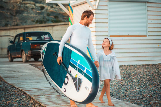 Smiling father and daughter walking together to surfing with sup board. Summer family vacation