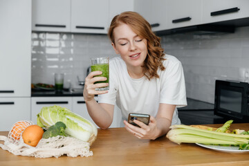 Young caucasian woman drinking fresh green smoothie uses smartphone to count calories in kitchen