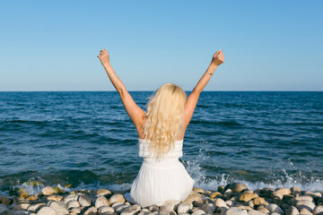 Blonde woman sitting on a pebble beach, arms raised.