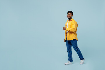 Full body young man of African American ethnicity 20s wear yellow shirt walking going strolling look camera isolated on plain pastel light blue background studio portrait. People lifestyle concept