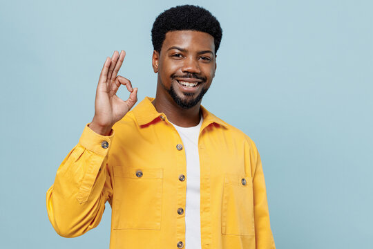 Young satisfied cheerful happy man of African American ethnicity 20s wear yellow shirt showing okay ok gesture isolated on plain pastel light blue background studio portrait. People lifestyle concept.