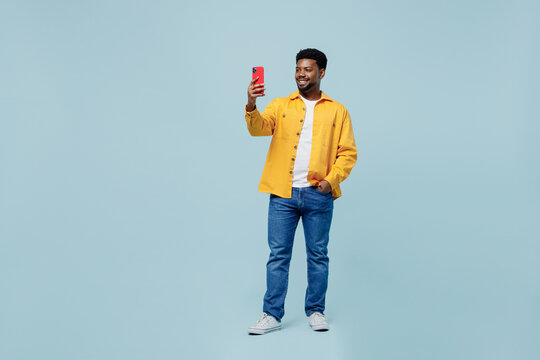 Full body young man of African American ethnicity wear yellow shirt doing selfie shot on mobile cell phone post photo on social network isolated on plain pastel light blue background studio portrait