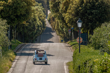 An old light blue Fiat 500 convertible with a suitcase on the back, drives along a tree-lined avenue of Tuscany, Artimino, Italy - obrazy, fototapety, plakaty