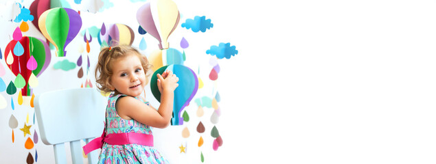 Caucasian curly haired girl in a beautiful blue dress on a white isolated background sits on a highchair. Multi-colored balloons, raindrop made of paper. Education, creativity, child care. Copy space